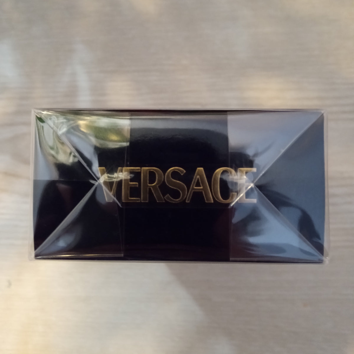 A black Versace box with gold lettering, sealed in clear plastic, placed on a light-colored wooden surface.