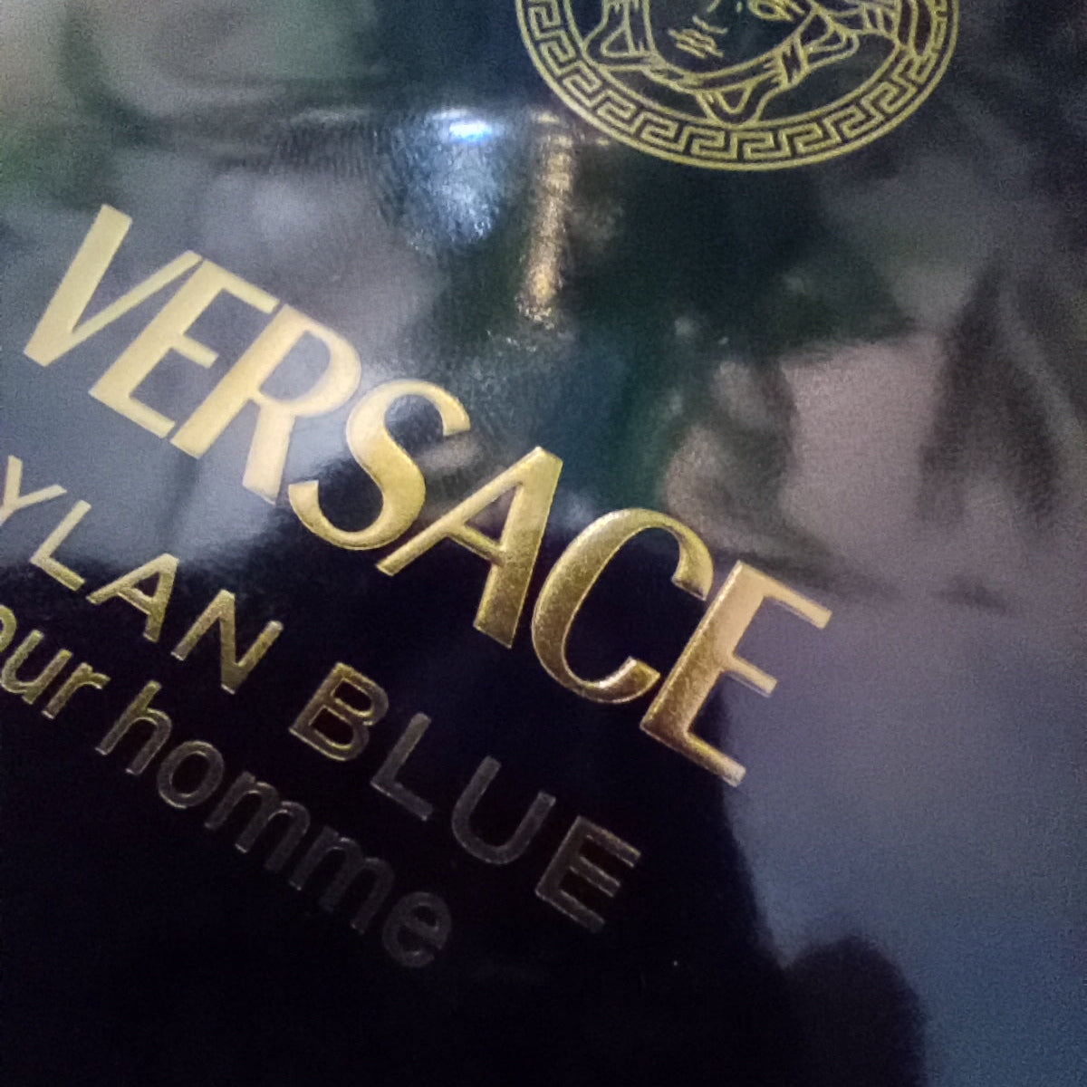 Close-up of a Versace makeup package showing the gold Versace logo, Medusa emblem, and text reading 'CYAN BLUE' and 'pour homme' on a glossy dark background.