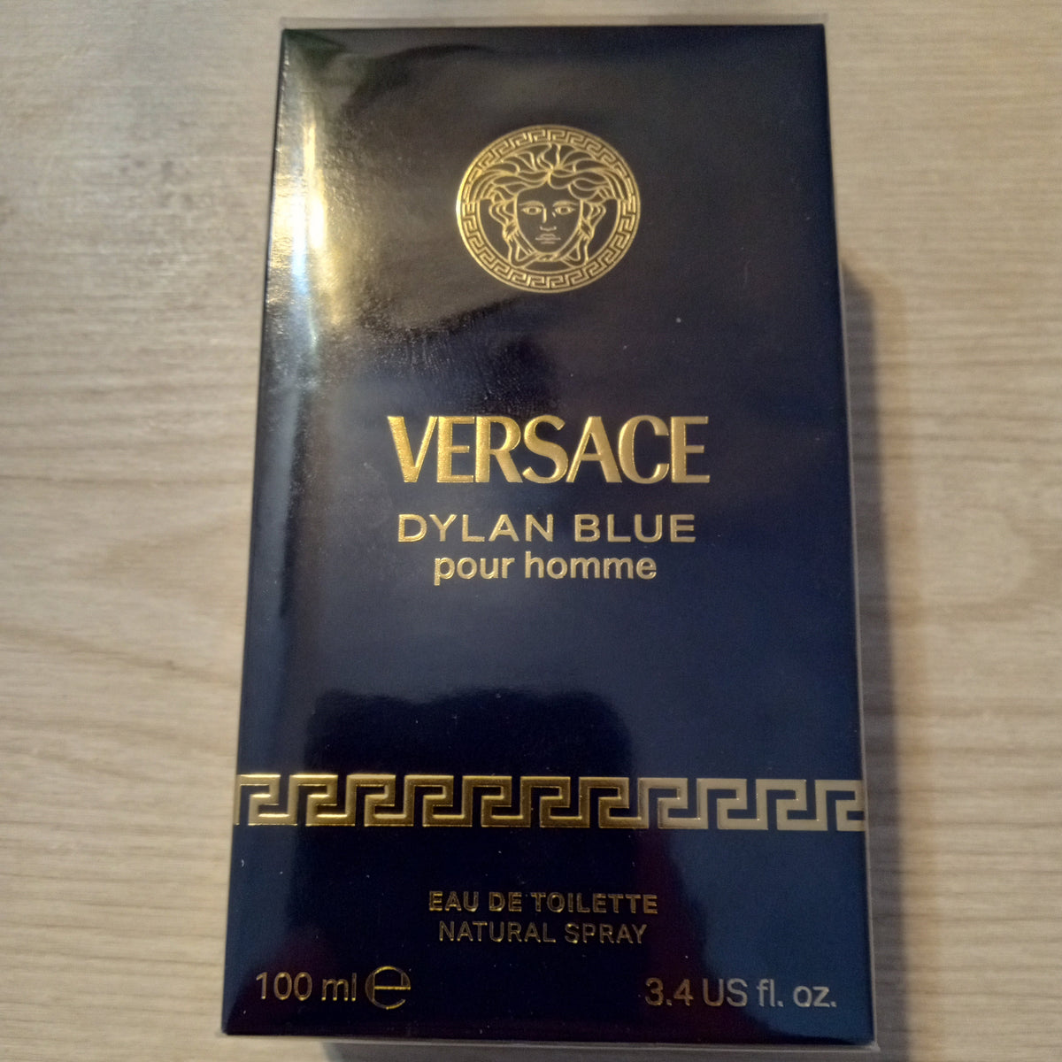 A 100ml box of Versace Dylan Blue pour homme Eau de Toilette, featuring a dark navy blue box with gold lettering and the iconic Medusa logo, placed on a light wood surface.