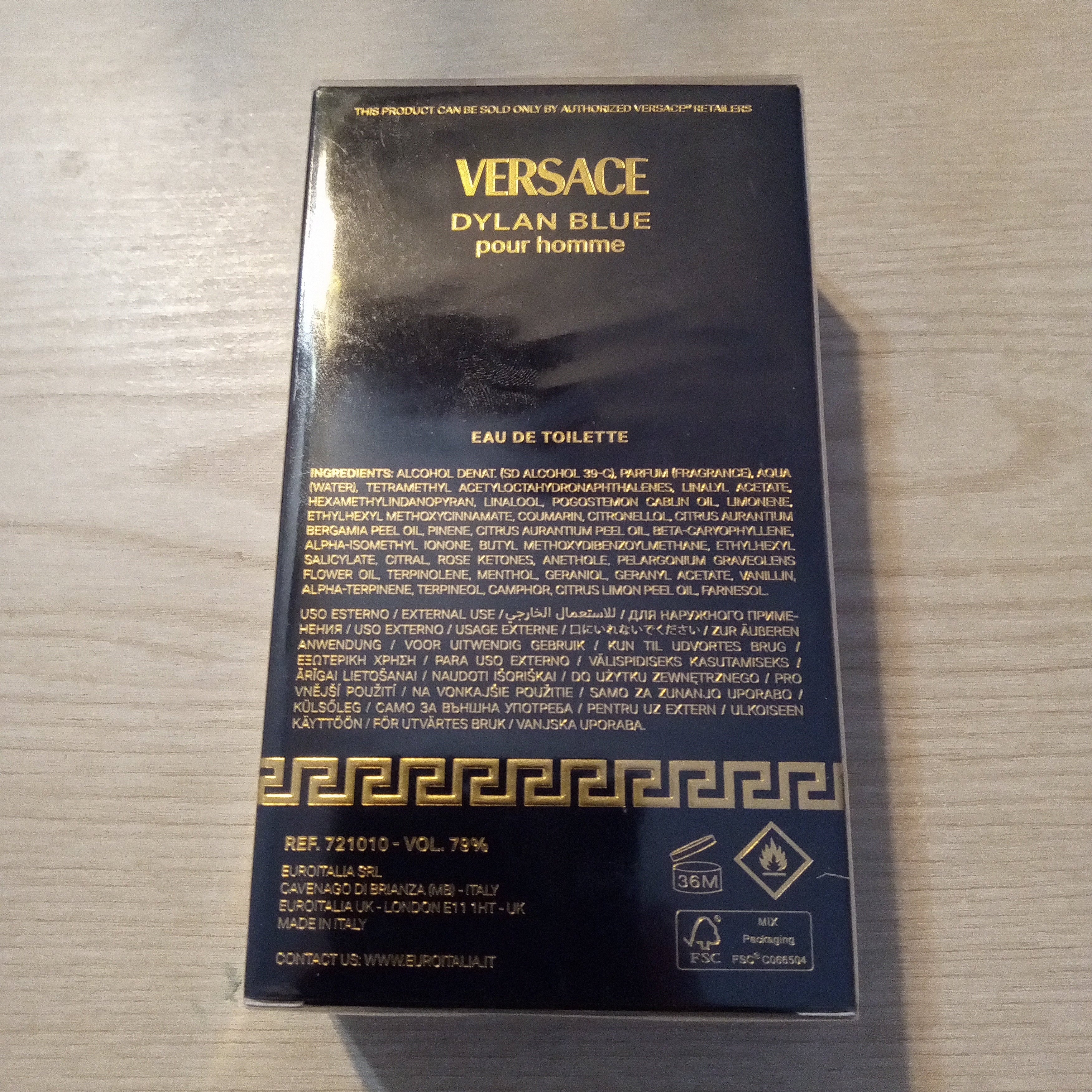 A black Versace Dylan Blue pour Homme Eau de Toilette box with gold lettering, photographed on a light wood surface.