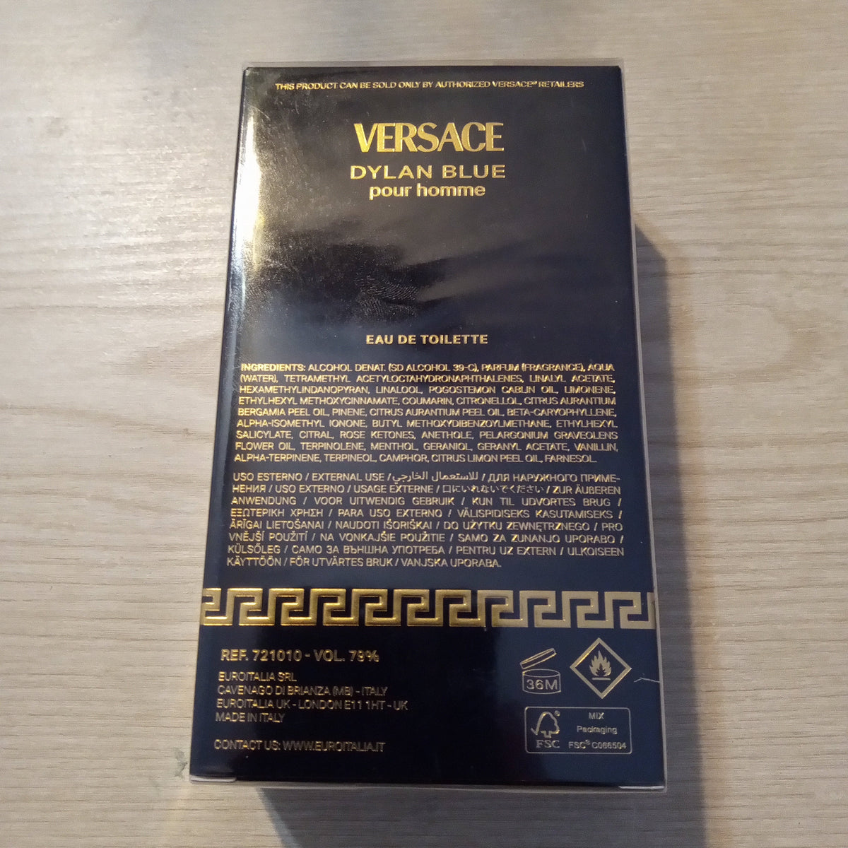 A black Versace Dylan Blue pour Homme Eau de Toilette box with gold lettering, photographed on a light wood surface.