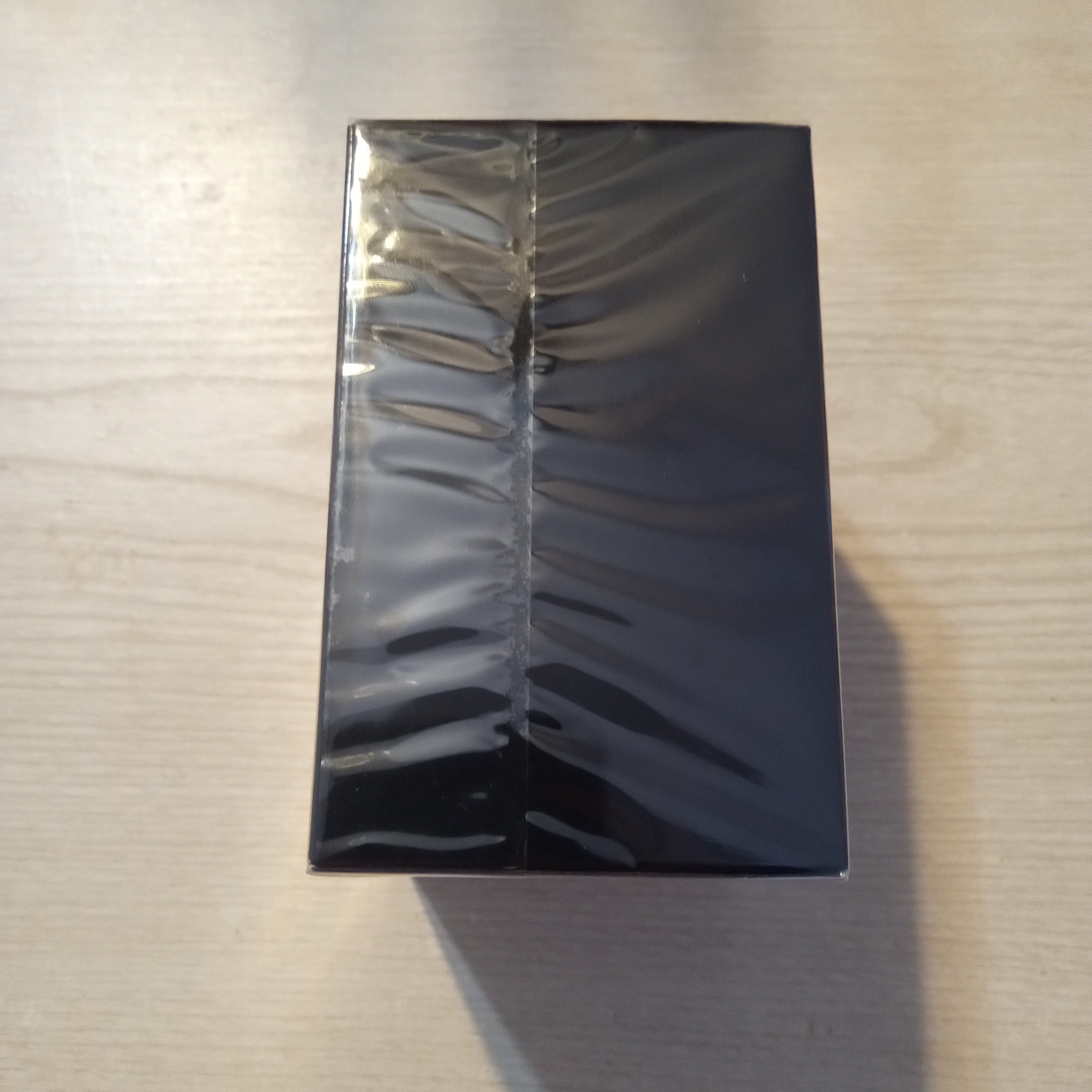 A black rectangular box wrapped in clear plastic, placed on a light wood surface with a red background visible at the top and bottom edges.