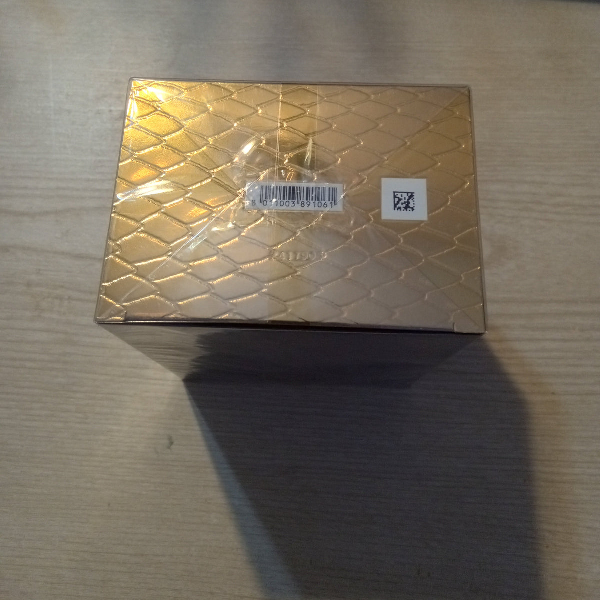 A square metallic gold box with a textured reptile pattern, sealed in clear plastic, sitting on a light-colored wooden table.