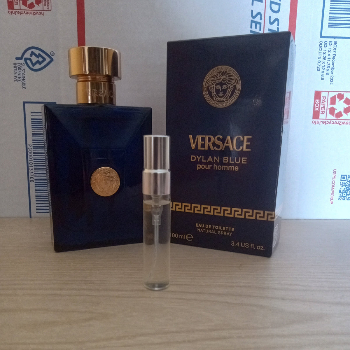 A Versace Dylan Blue Pour Homme eau de toilette sample vial with a silver cap stands in front of its dark blue box and the original bottle, placed on a light wood surface.