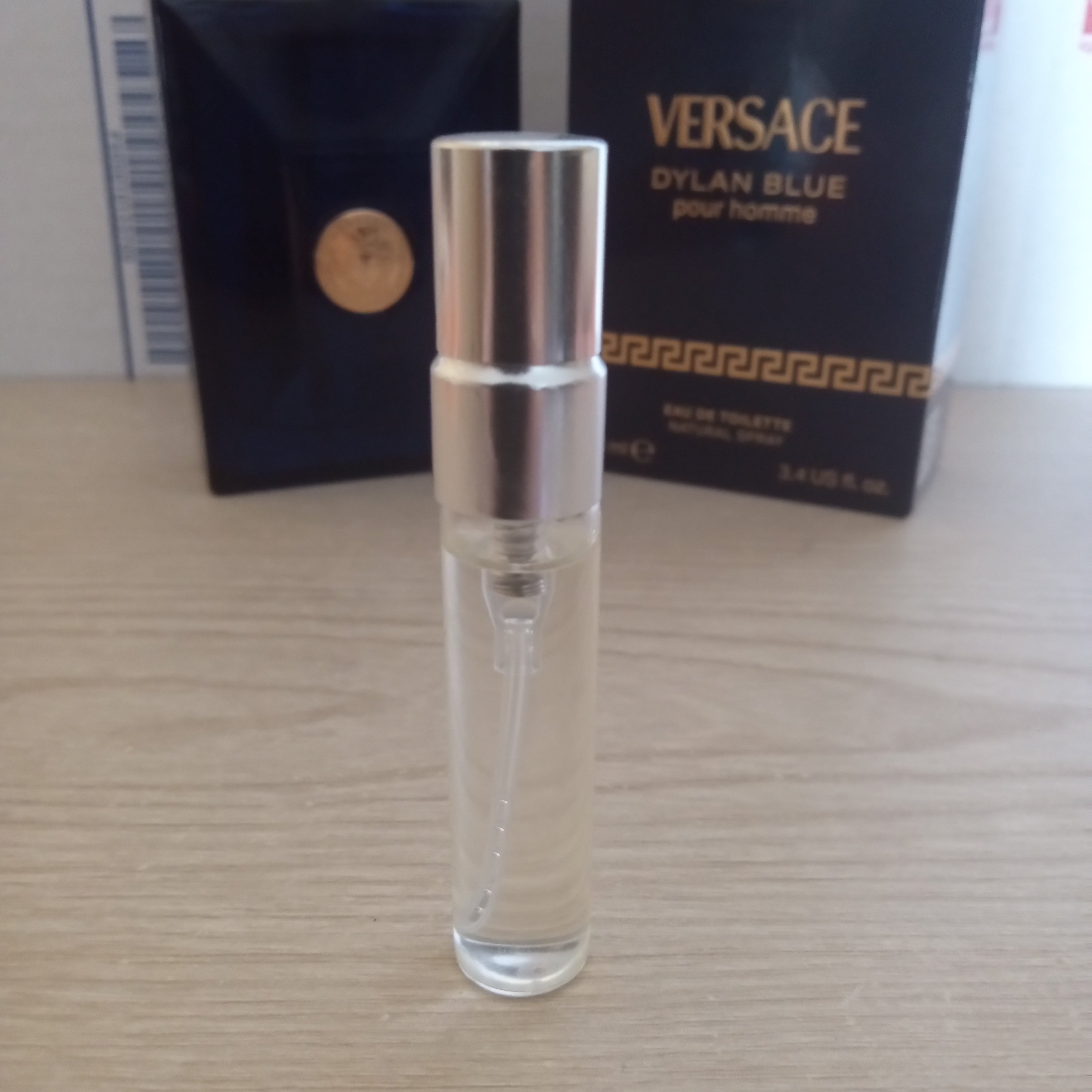 A small clear Versace Dylan Blue for grooming perfume spray bottle with a silver cap, standing on a light wood surface with the original black and gold Versace box in the background.