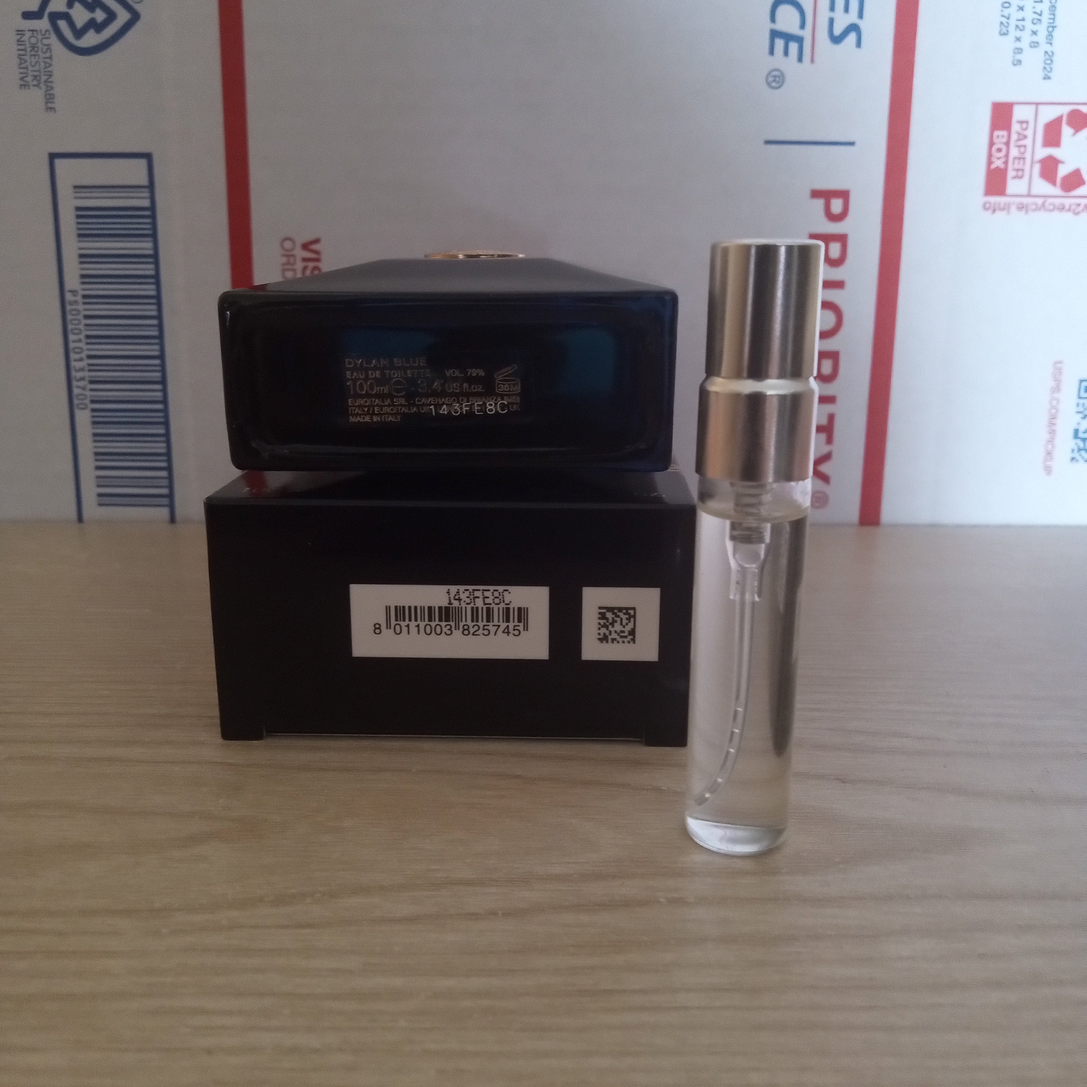 A small clear glass perfume sample spray bottle with a silver atomizer cap stands next to its black retail box on a light wood tabletop, with a USPS shipping box in the background.