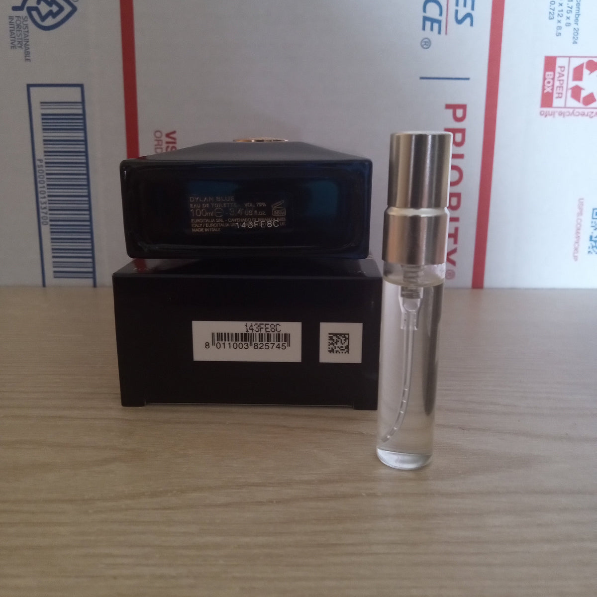A small clear glass perfume sample spray bottle with a silver atomizer cap stands next to its black retail box on a light wood tabletop, with a USPS shipping box in the background.