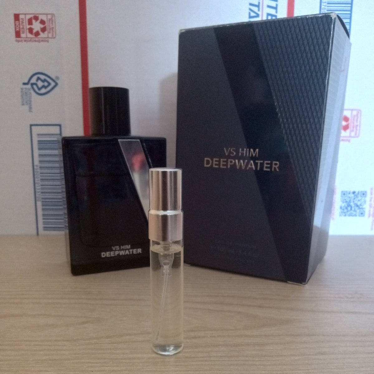 A small clear glass perfume sample vial with a silver cap stands in front of a black VS HIM DEEPWATER bottle and its matching black box on a light wood surface.
