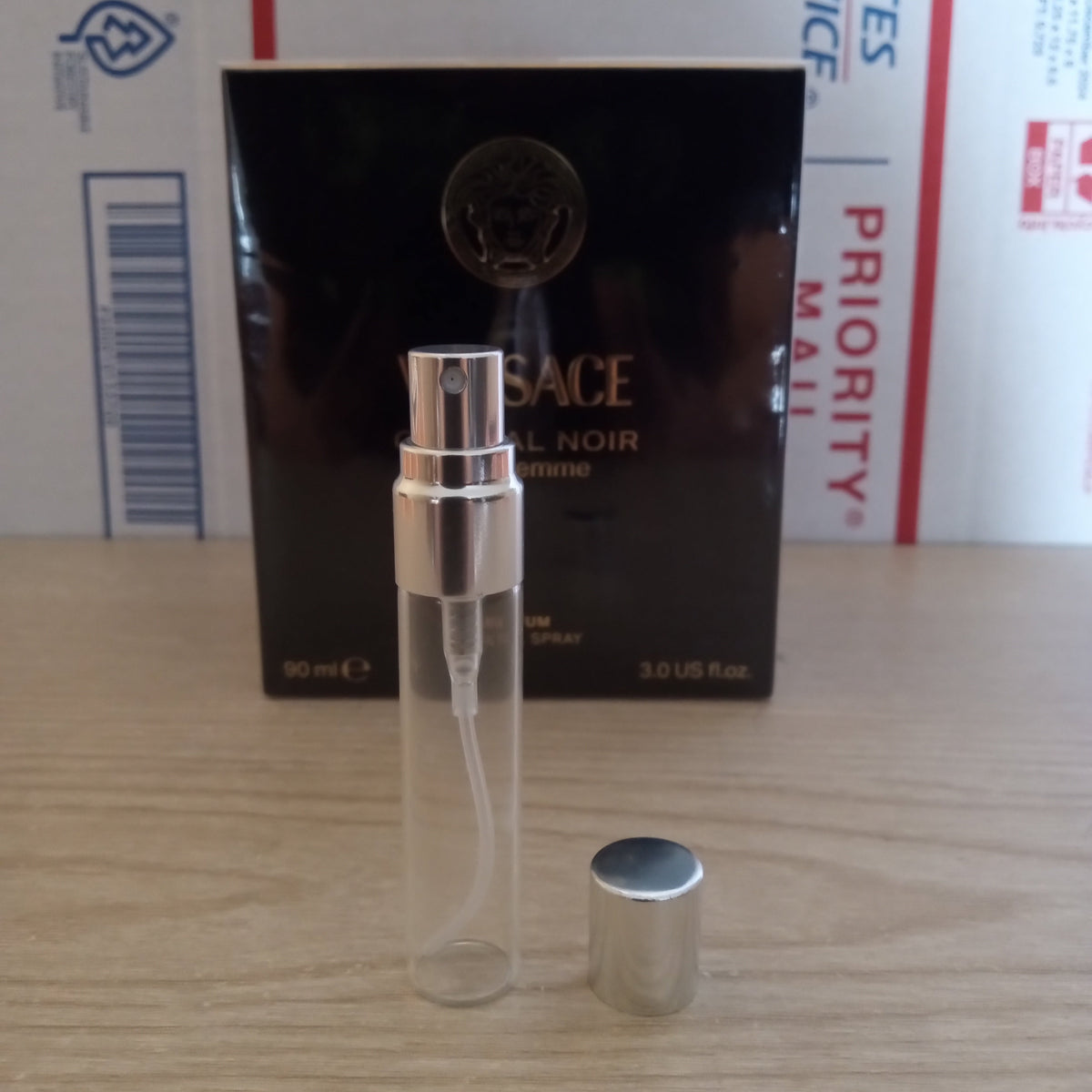 A Versace Elixir Noir 10ml clear glass perfume atomizer spray bottle with a silver cap, shown next to its black retail box on a wooden tabletop with shipping boxes in the background.