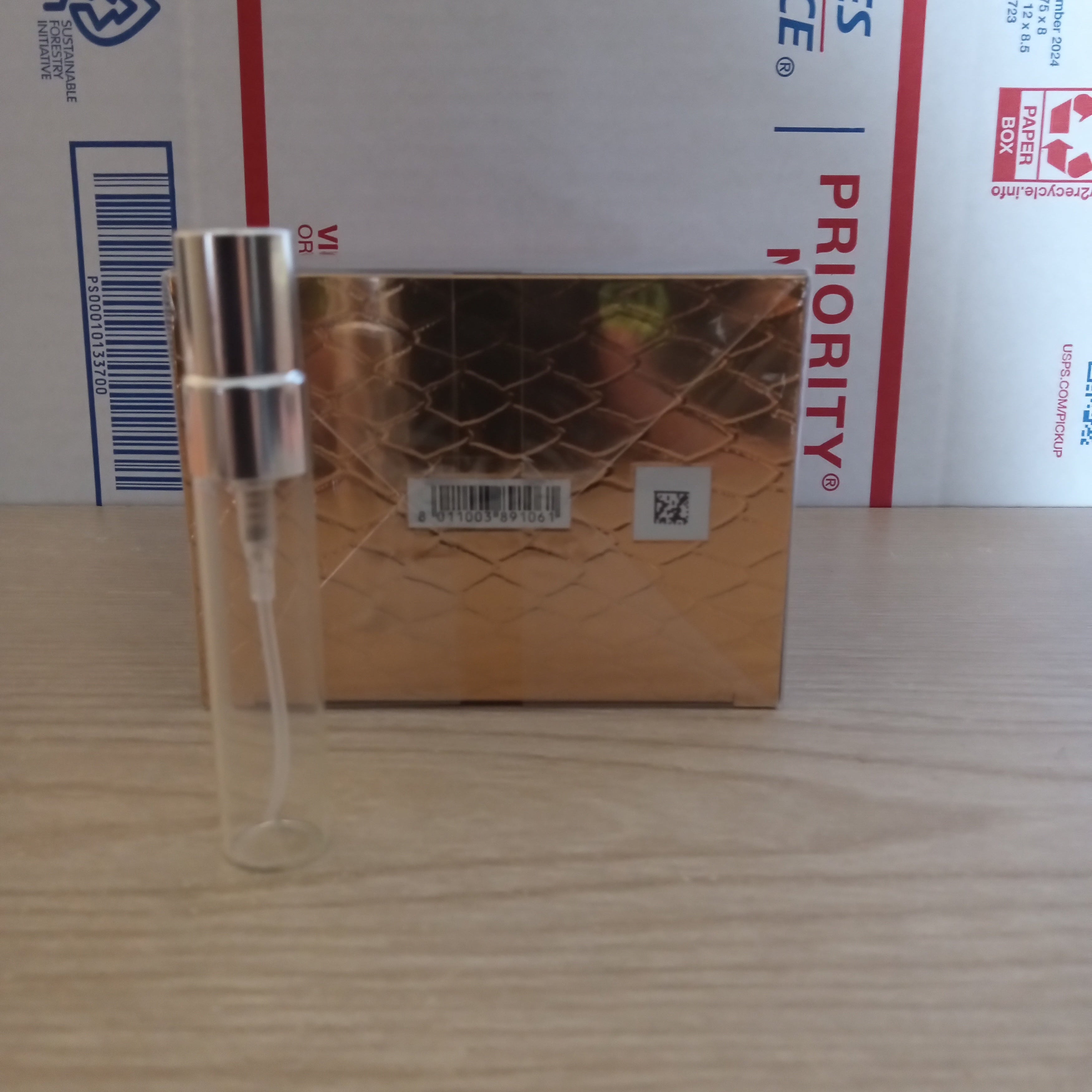 A small clear glass spray bottle with a silver atomizer cap resting on top of a gold rectangular box, placed against a white USPS Priority mailer box on a light wood surface.