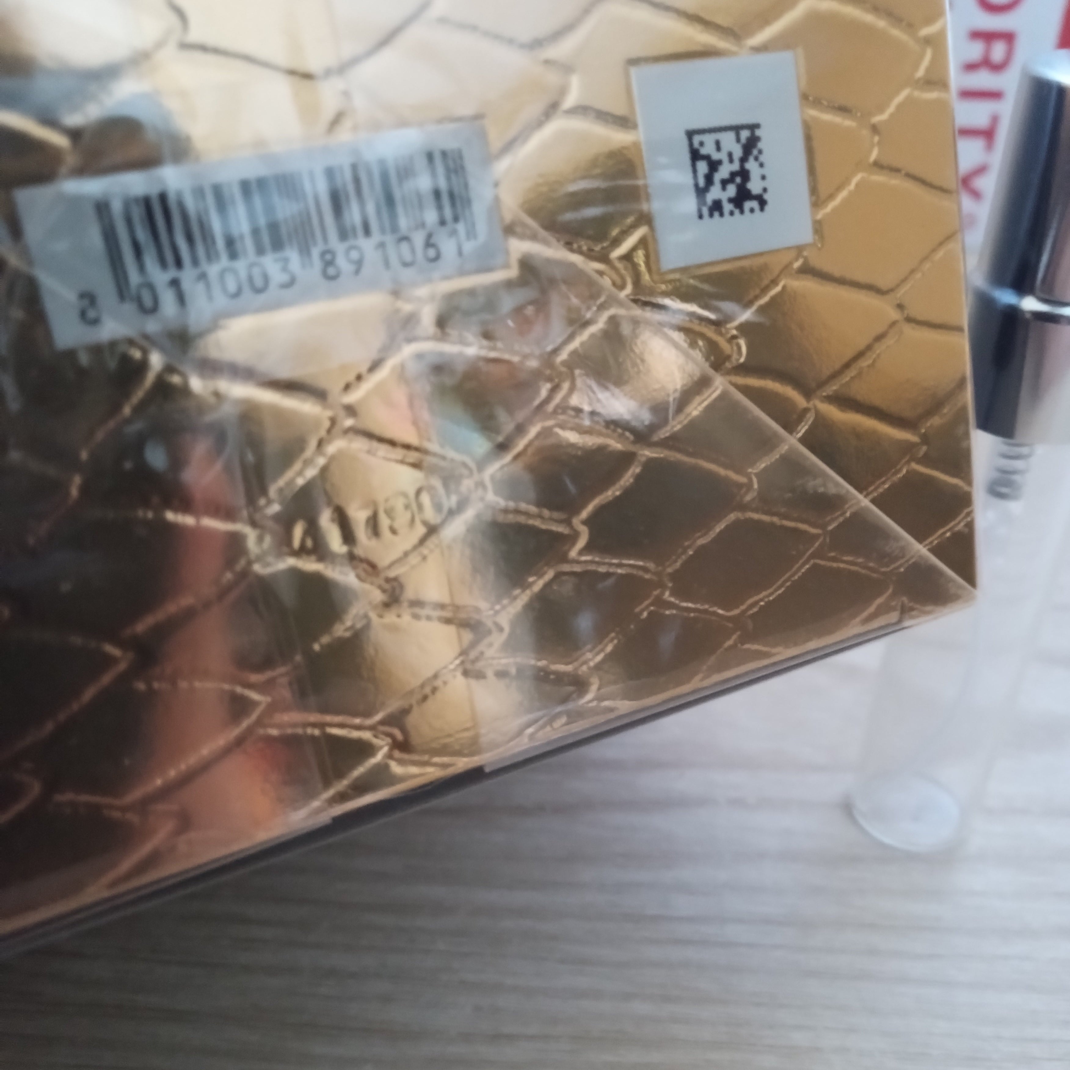 A close-up of a gold-toned textured cosmetic compact or box with a diamond pattern, sealed in plastic with a barcode sticker and QR code label, placed on a light wood surface.